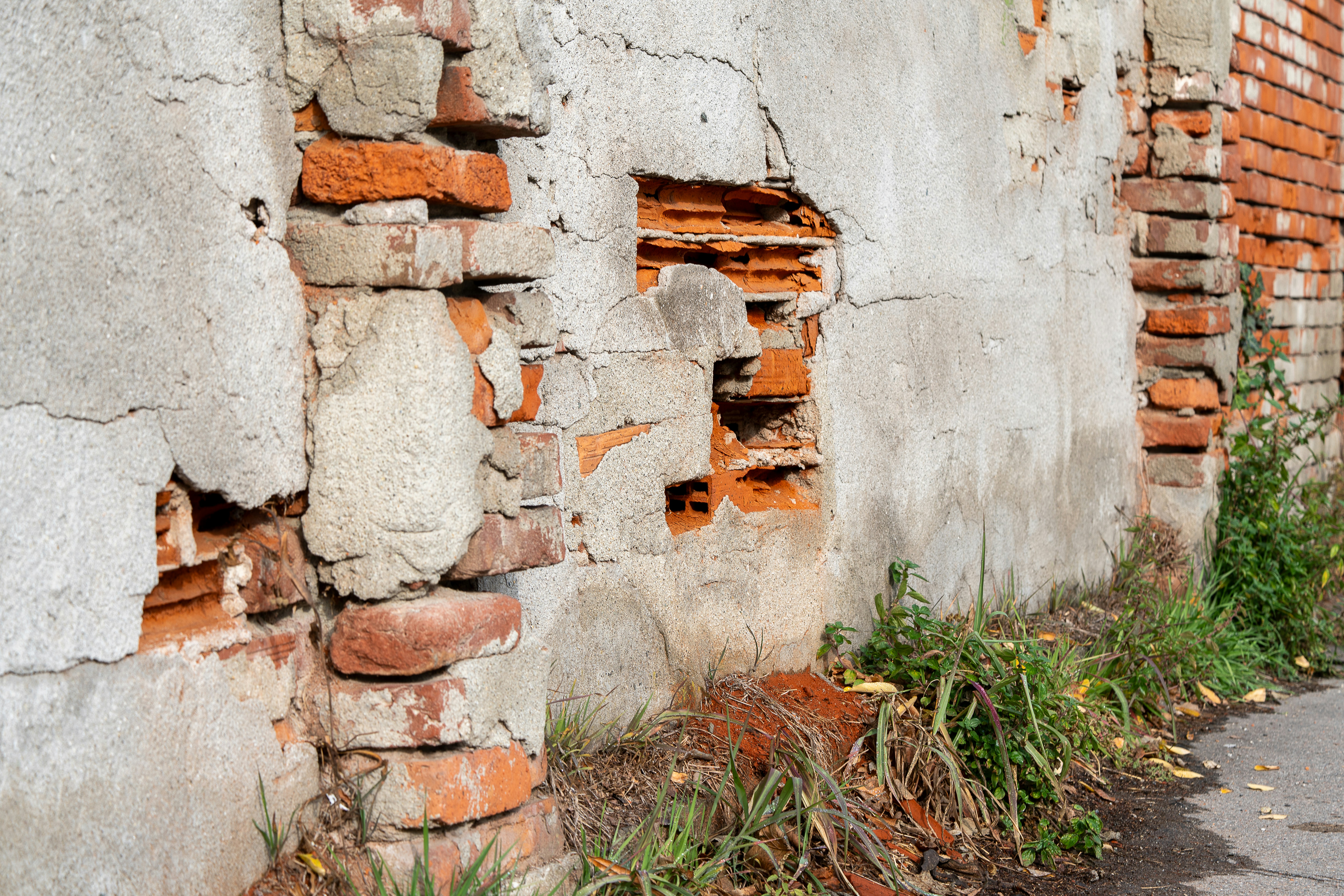 Damaged brick wall with crumbling plaster and overgrown weeds